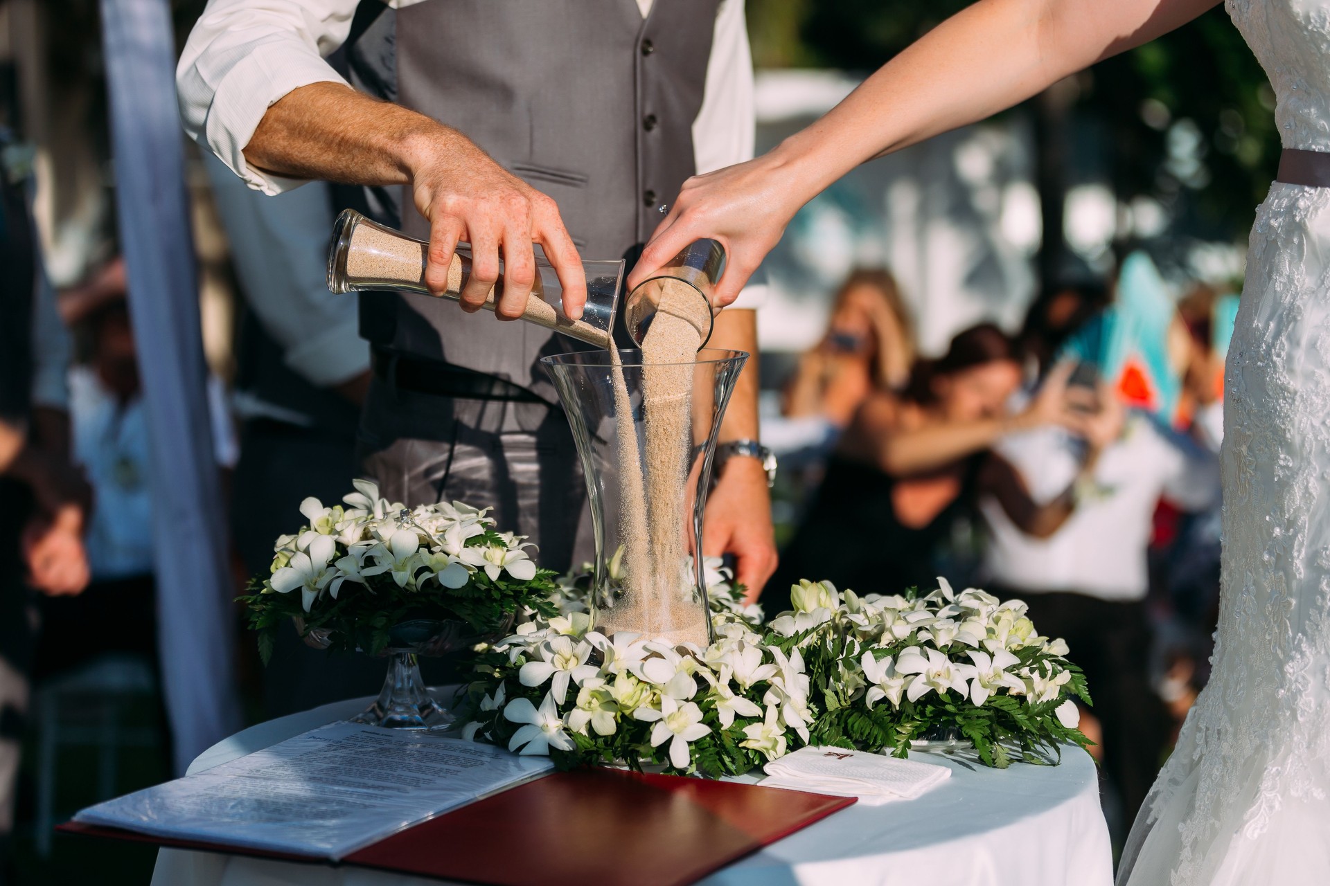 The bride and groom are pouring sand together at a wedding ceremony at a hotel. On the table was a beautiful bouquet of white flowers. There were many guests in attending.